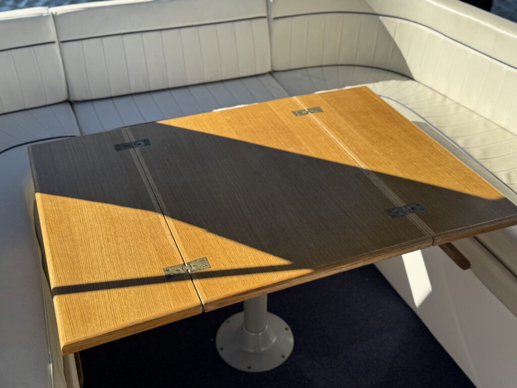 A rectangular teak boat table with a dark central inlay sits in a cream upholstered seating area, with sunlight casting a diagonal shadow across the surface.
