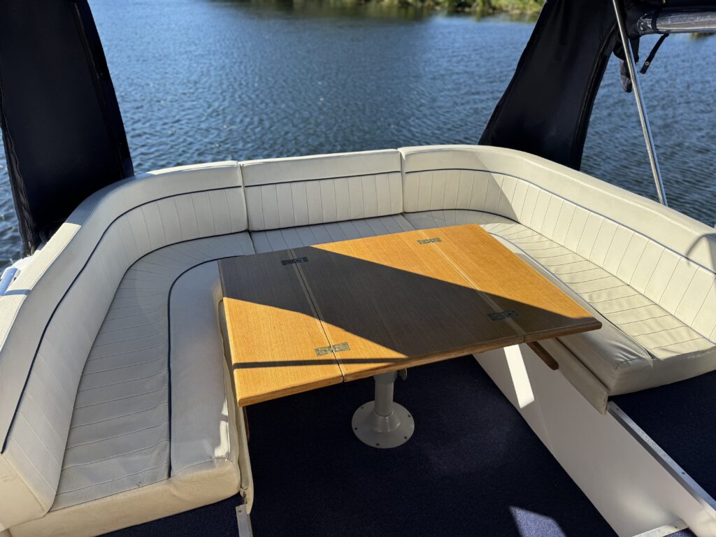 A U-shaped cream upholstered seating area on a boat surrounds a light wood table with a dark center panel, with blue water visible beyond the deck.