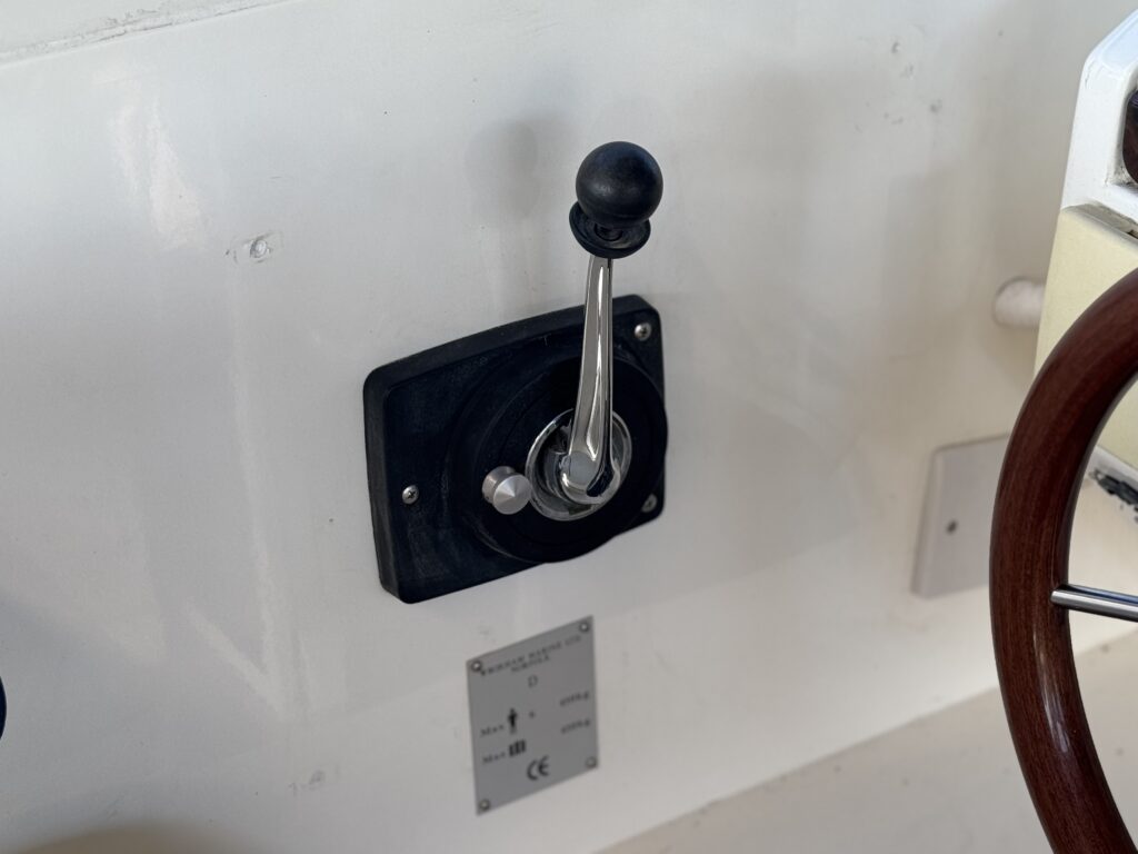 A chrome lever control with a black knob is mounted on a white fiberglass surface beside a wooden steering wheel and a small metal identification plate.