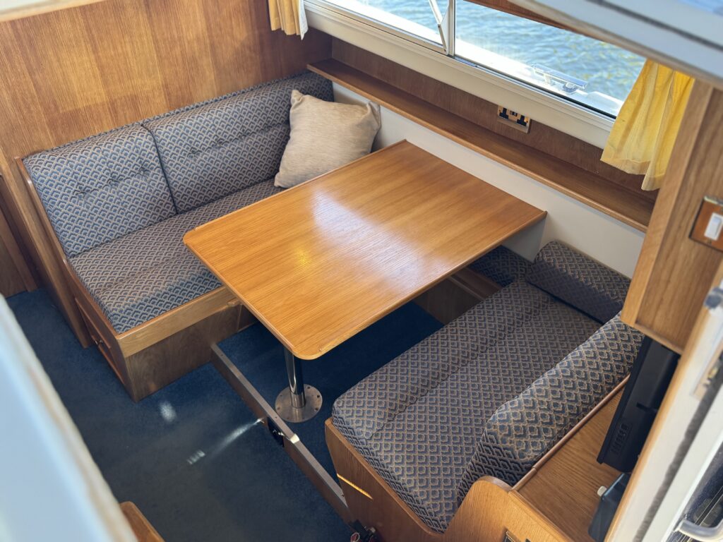A compact boat cabin corner with two patterned bench seats around a wooden table, blue carpet, wood-paneled walls, and a window with yellow curtains.