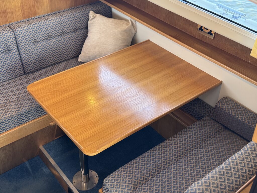 A rectangular wooden table is set between upholstered bench seats in a compact cabin, with a window and beige cushion above the seating.