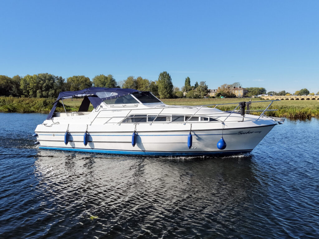 A white motor yacht with a blue canopy and fenders floats on calm water, with a tree-lined shore and clear blue sky in the background.