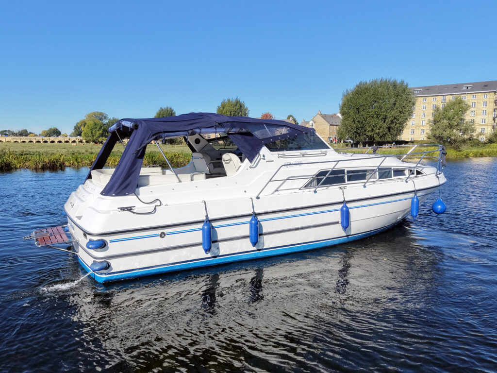 A white motor yacht with a dark blue canopy and blue fenders floats on calm water near grassy banks and buildings under a clear sky.