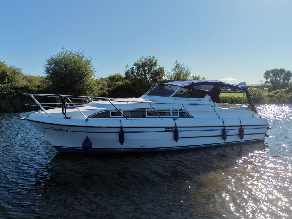 A white motor yacht with blue fenders floats on calm water under a clear blue sky, with trees and shoreline in the background.
