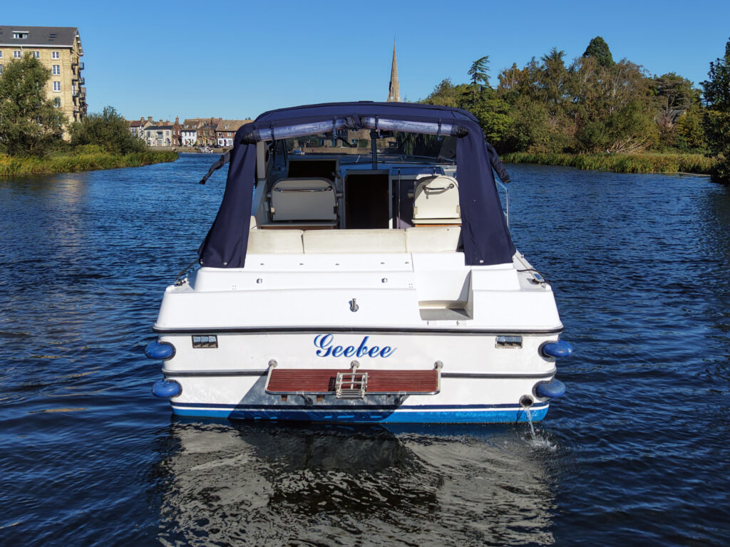 A white motorboat named Seebee moves on a river, with a blue canopy and trees lining the banks.